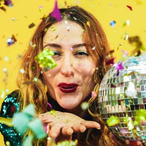 Happy fashion girl blowing confetti from hands holding a disco ball with yellow background